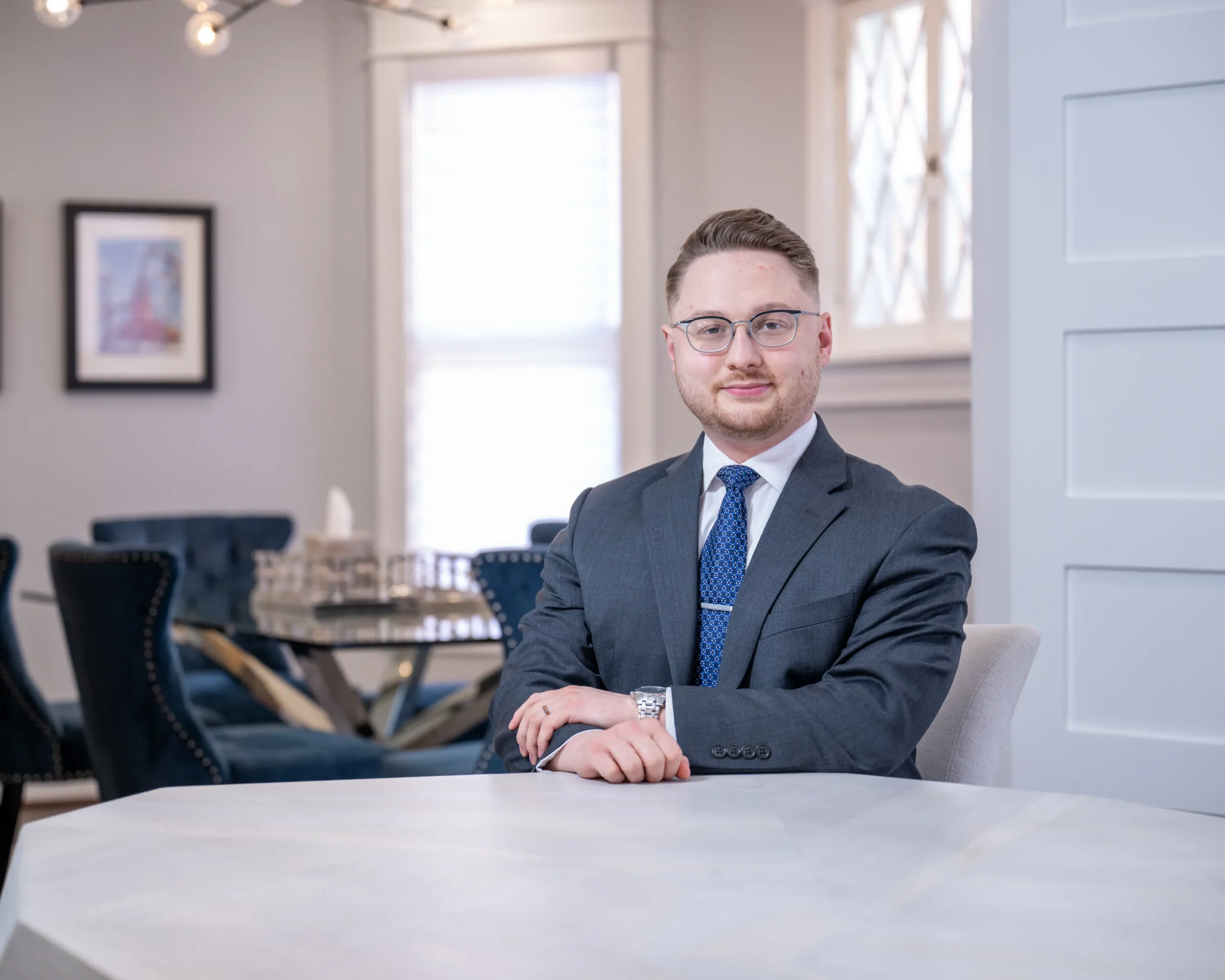 Professional man with glasses and suit seated at a marble table in an office setting.