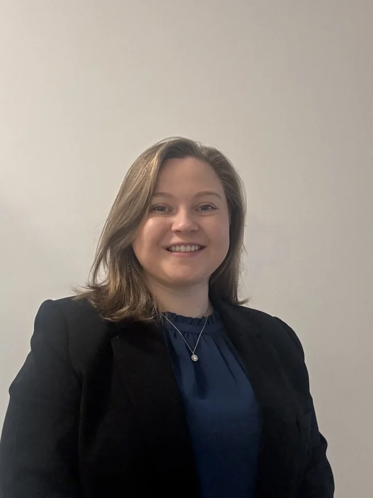 Professional woman with shoulder-length light brown hair, wearing a black blazer and blue blouse, smiling against plain background.