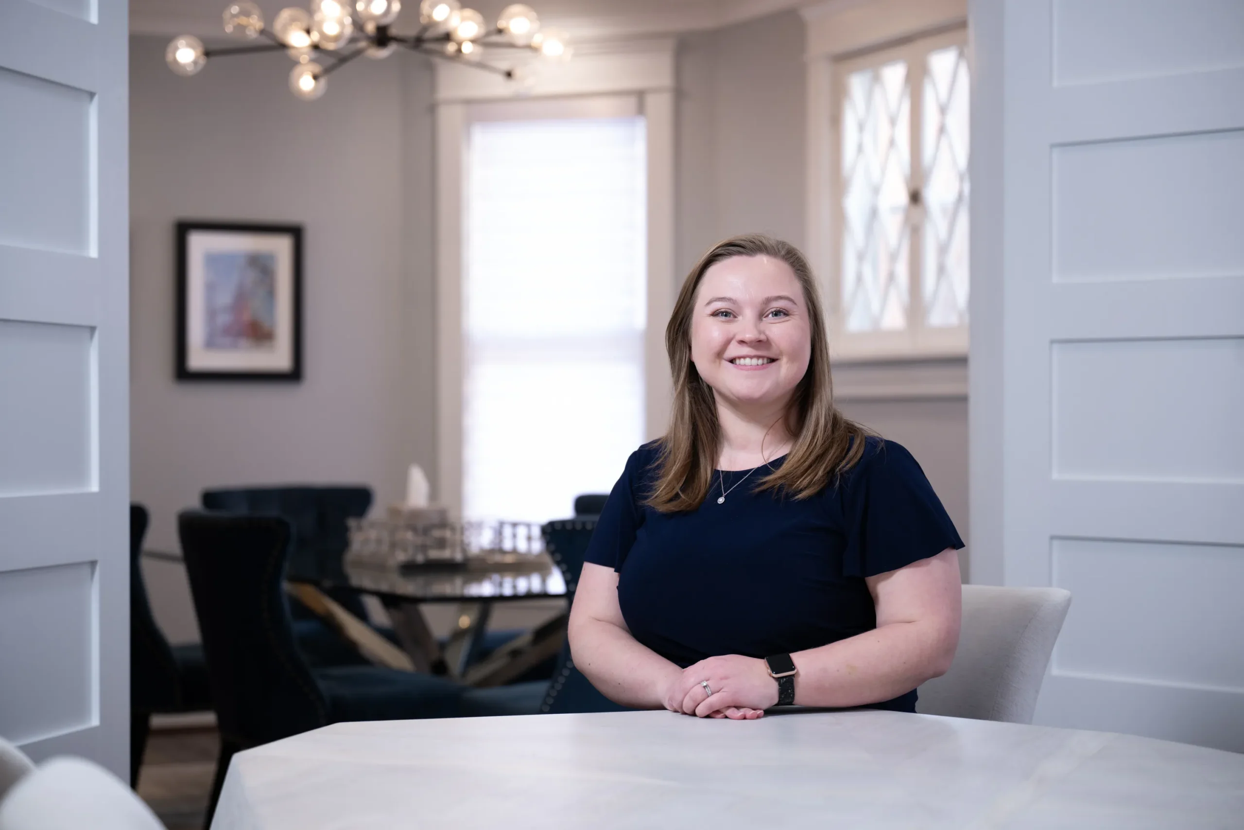 Smiling woman with light brown hair, navy blouse, and smartwatch seated indoors at white table.