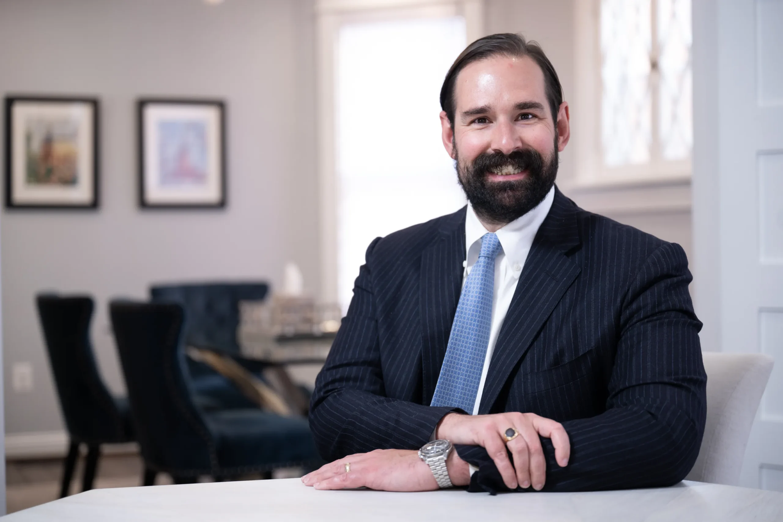 Professional man with beard and mustache in dark pinstripe suit and blue tie smiling, seated indoors.