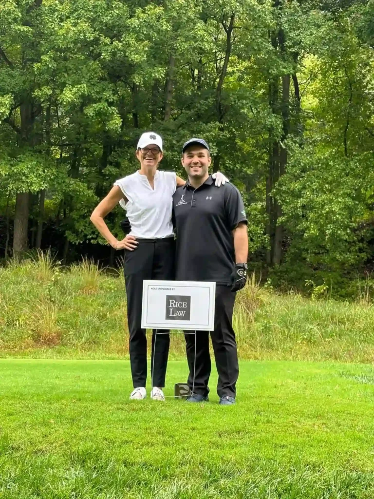 Two individuals smiling and posing together on a sunny golf course, surrounded by green grass and trees.