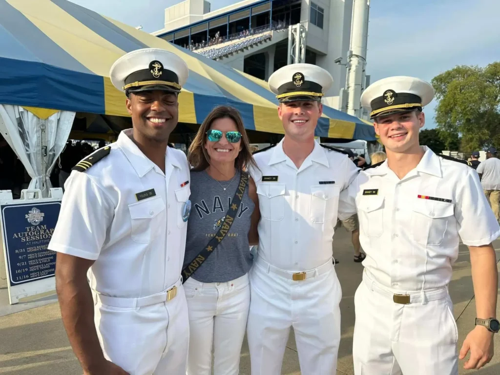 Three navy members smile for a photo with a woman dressed in white, standing together against a scenic backdrop.