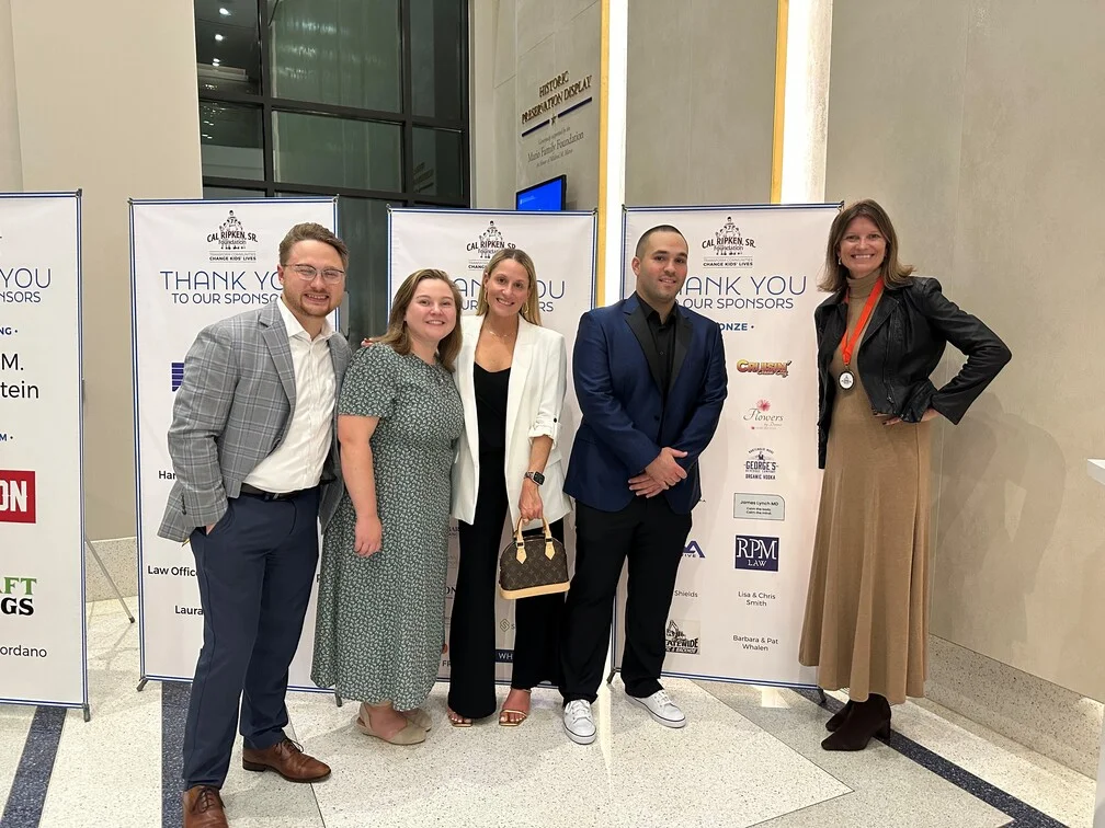A diverse group of people stands together in front of a large "Thank You" banner, smiling and celebrating.