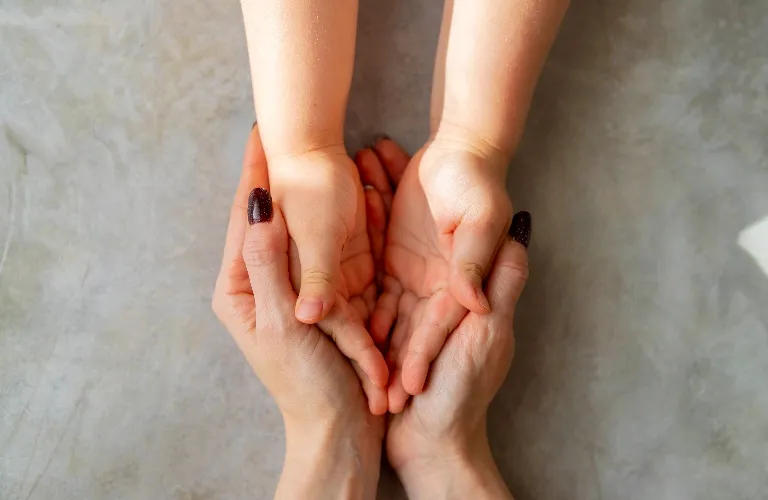 Close-up of an adult's hands gently holding a child's hands against a neutral background.