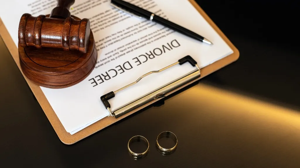 Wooden judge gavel, divorce decree document on clipboard, black pen, and two gold wedding rings on black surface.