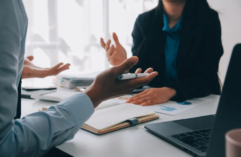 Two professionals engaged in a discussion at a desk with documents, a notebook, and a laptop.