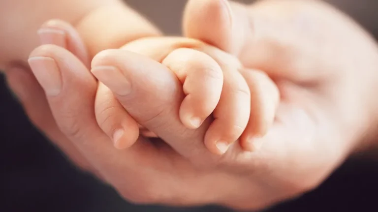 Close-up of an adult hand gently holding a newborn baby's hand with soft lighting.
