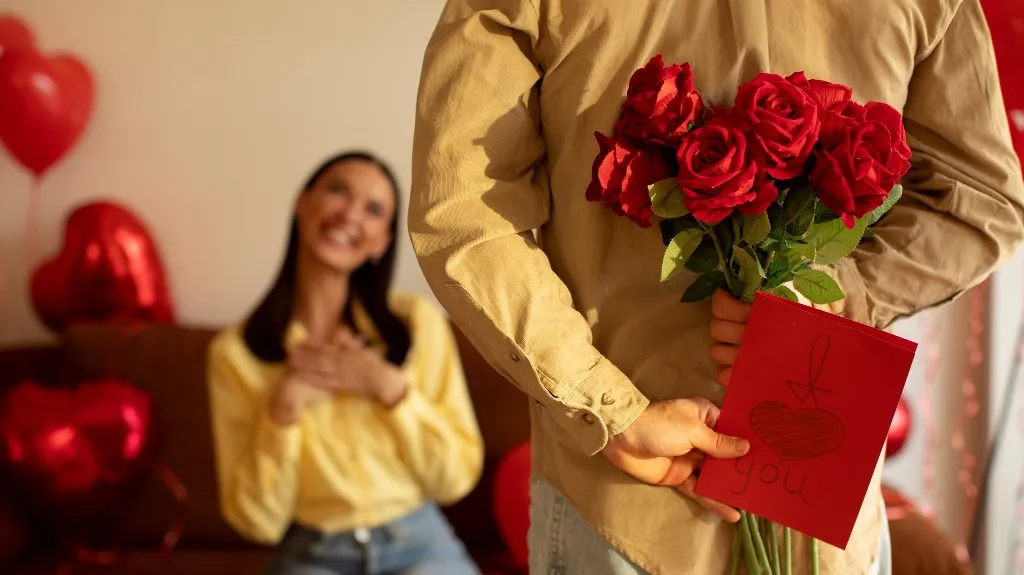 Man holding red roses and a handmade "I love you" card behind his back, woman smiling in background.