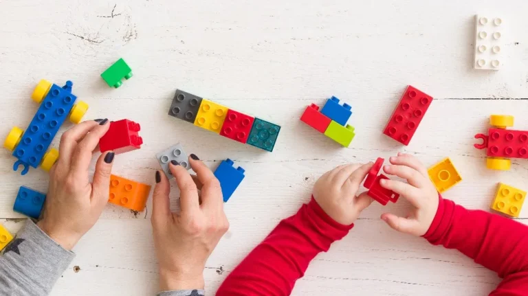Adult and child hands assembling colorful plastic building blocks on white surface.