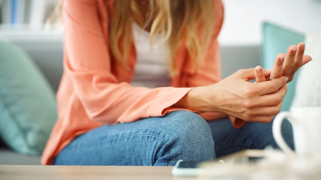 Woman in casual clothing sitting on a couch, holding her wrist, with a smartphone and a white cup on the table.