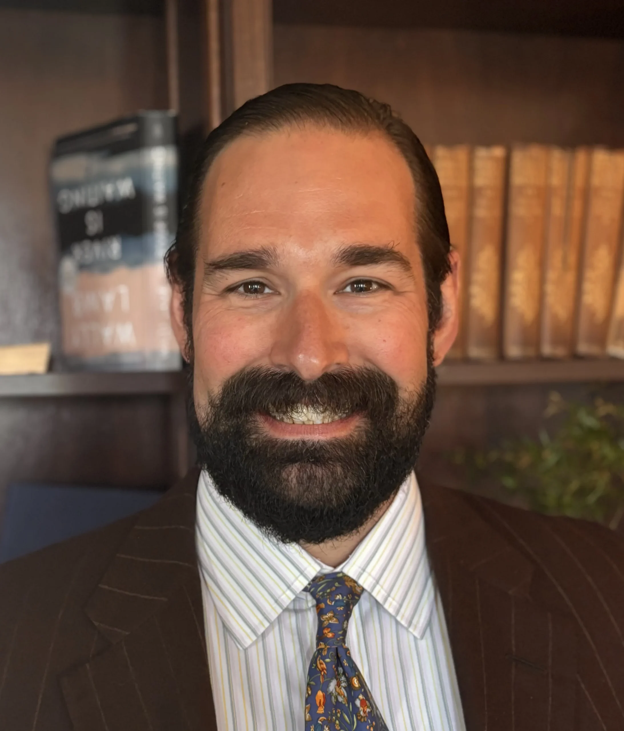 Professional man with dark hair and beard, wearing a striped shirt, floral tie, and brown suit jacket, smiling in front of a bookshelf.