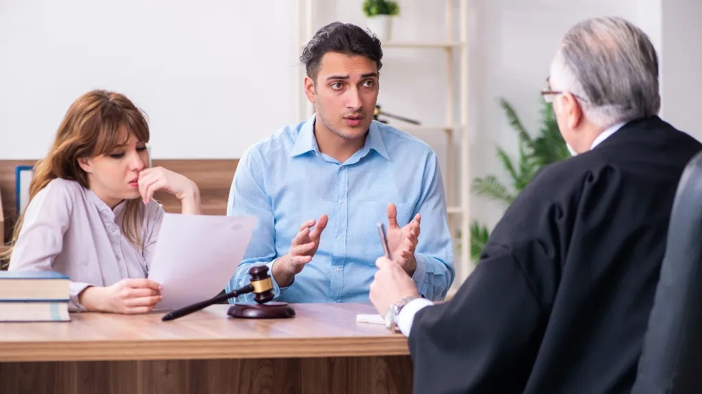 Three people in a legal consultation, including a man in a blue shirt, a woman reviewing documents, and a judge in a black robe.