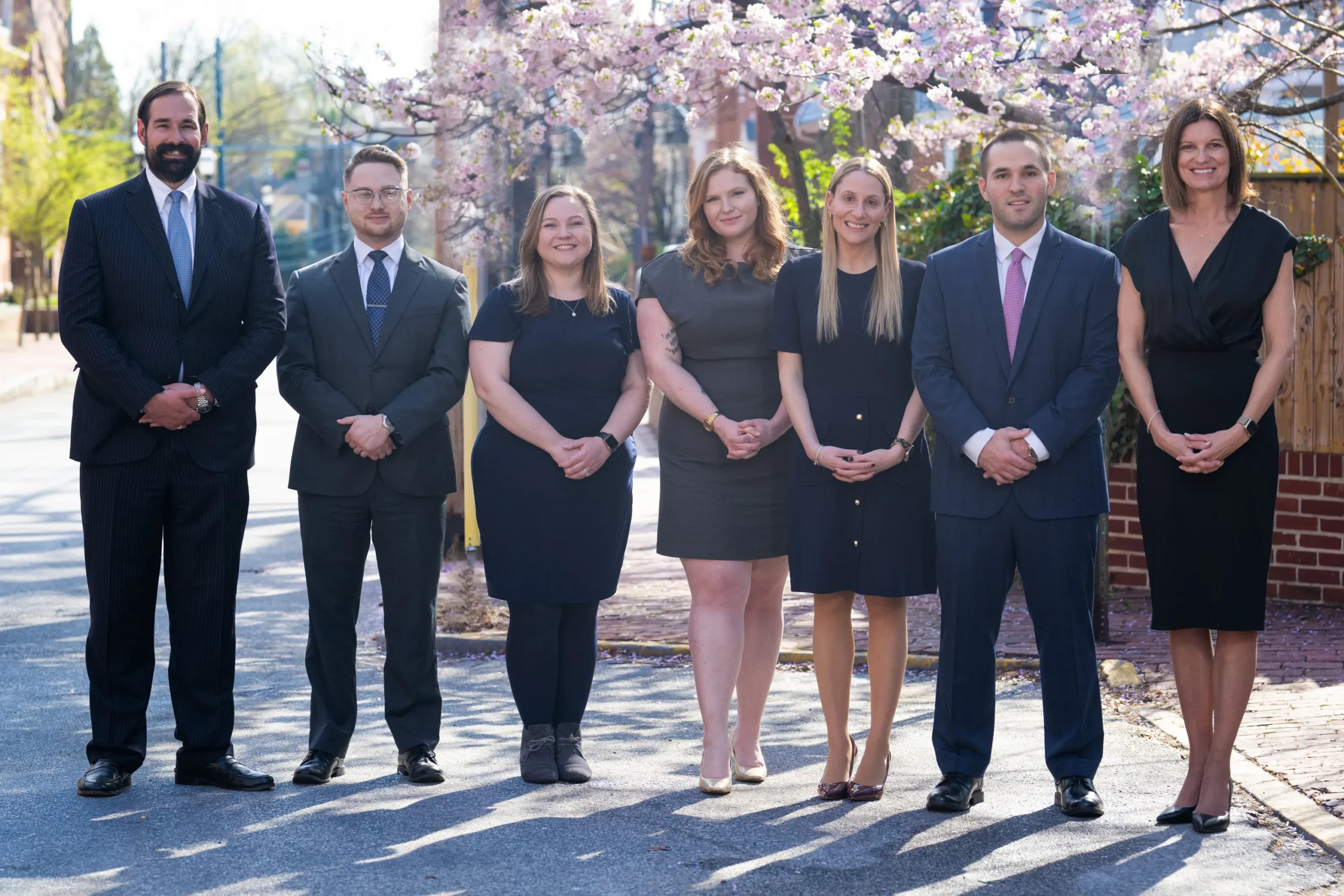 Group of seven professionally dressed men and women standing outdoors on a sunny day with cherry blossom trees in the background.