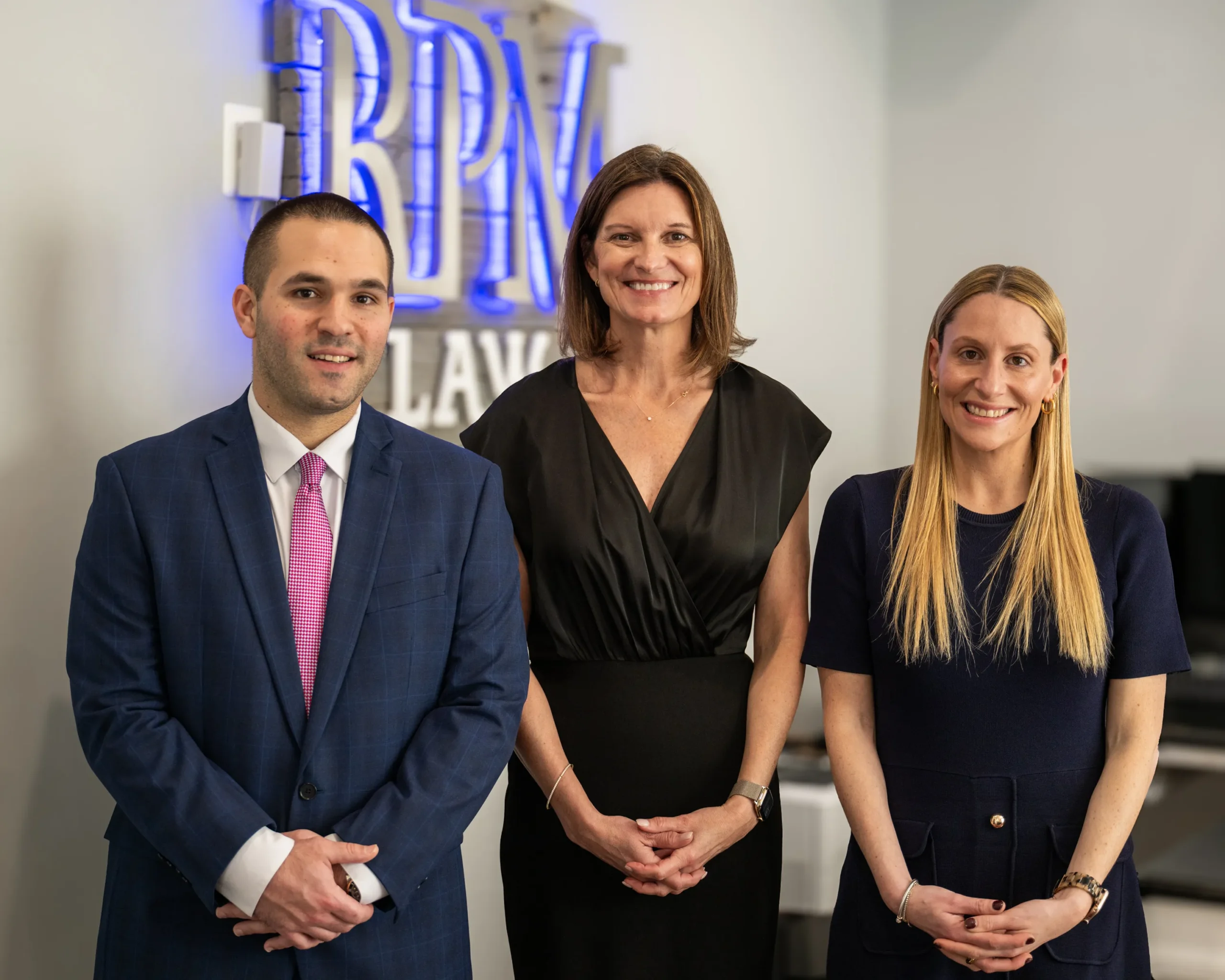 Three professionally dressed individuals standing indoors in front of a wall with a blue neon sign that reads "BRIAN LAW.