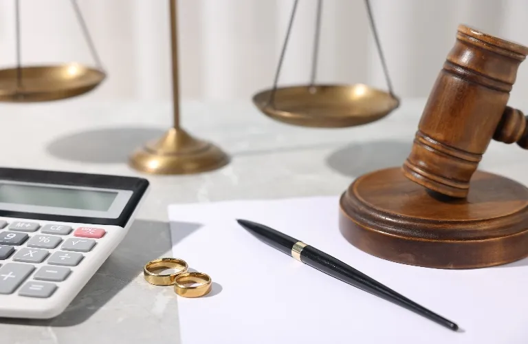Wooden judge's gavel, gold wedding rings, black pen, calculator, and brass balance scale on white surface.
