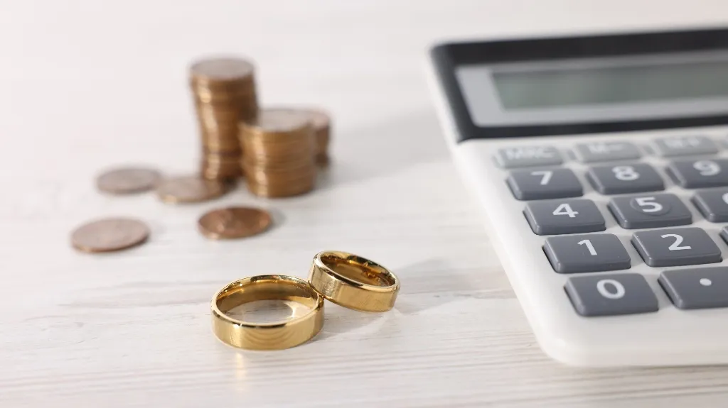 Two gold wedding rings, stacked coins, and a calculator on a white surface.