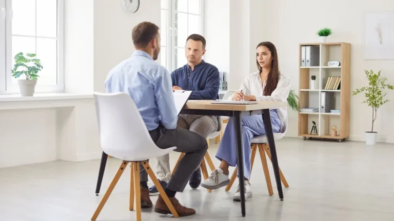 Three people engaged in a formal discussion around a wooden table in a bright office.