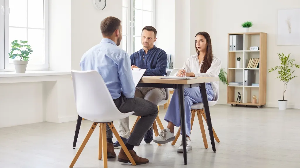 Three people engaged in a formal discussion around a wooden table in a bright office.