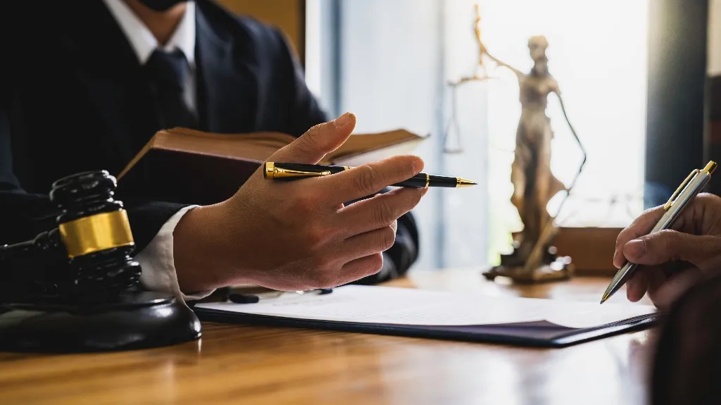 Two individuals in a legal consultation, one holding a pen and book, with a gavel and Lady Justice statue on the table.