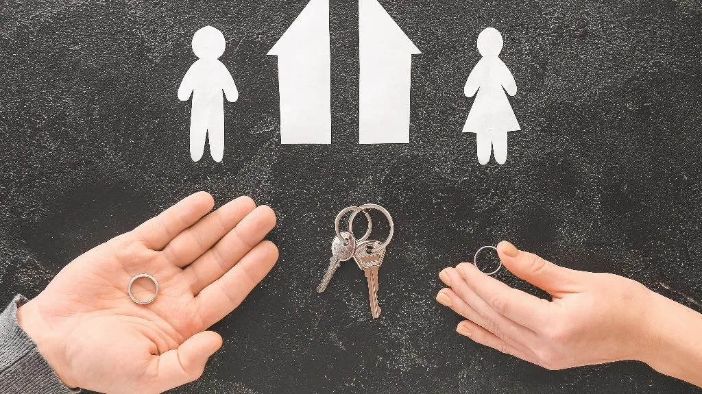 Two hands holding wedding rings below white paper cutouts of a man, house, and woman on black background.