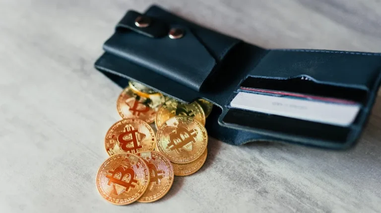 Black leather wallet with several gold Bitcoin coins spilling out, placed on a light surface.