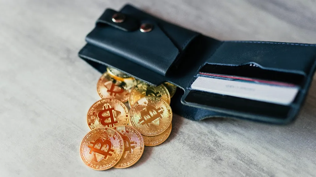 Black leather wallet with several gold Bitcoin coins spilling out, placed on a light surface.