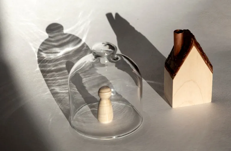 Wooden pawn figure under a glass dome beside a small wooden house model, casting shadows.