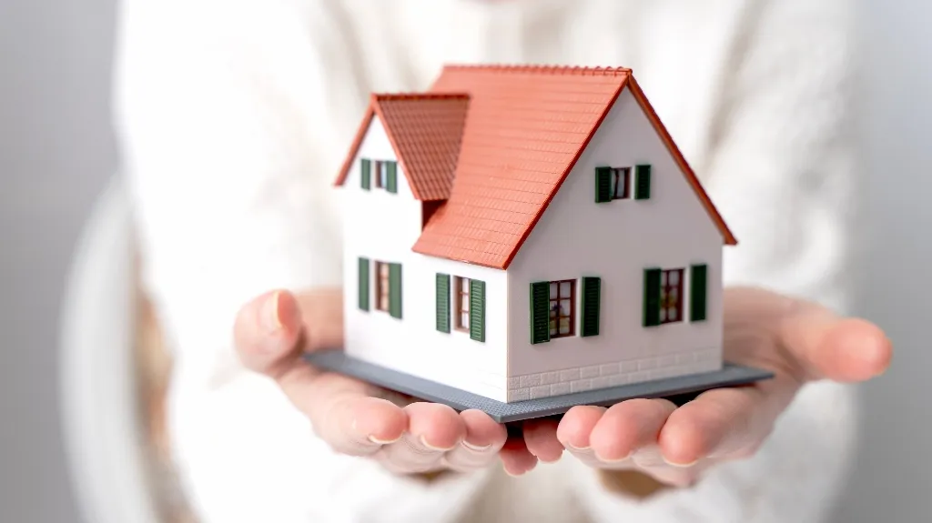 Hands holding a detailed miniature white house with red roof and green shutters.
