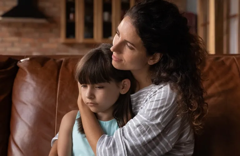 Woman with curly dark hair in a striped shirt comforting a sad young girl with dark hair on a brown leather couch.