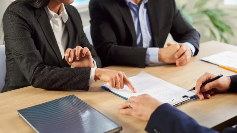 Three business professionals in suits discussing and reviewing documents at a wooden table.