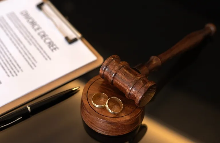 Wooden judge's gavel with two gold wedding rings, pen, and a blurred divorce document on clipboard.