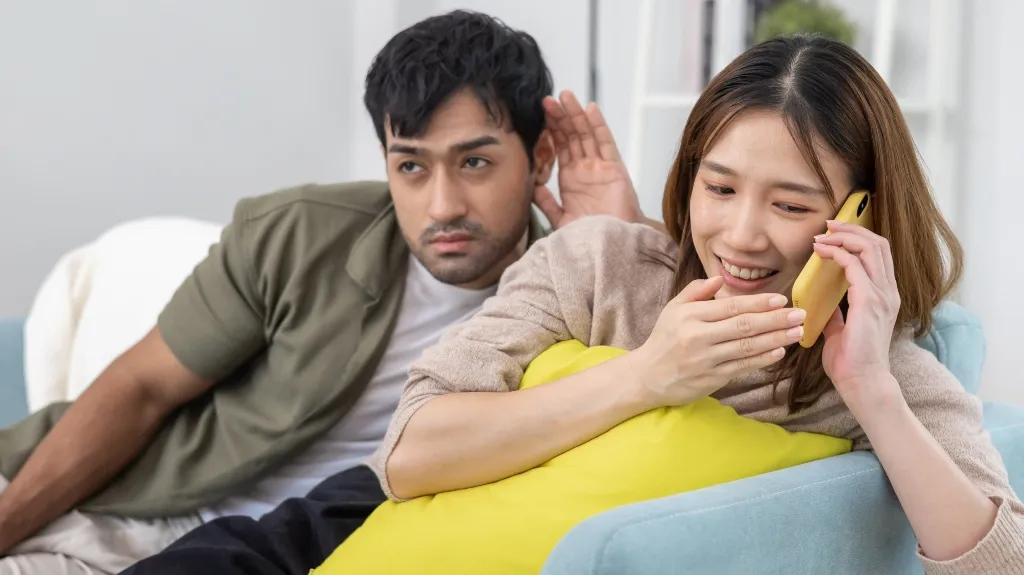 A woman smiling and talking on a yellow phone while a man listens intently beside her on a light blue couch.