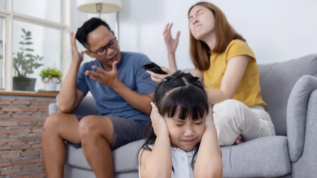 Young girl covering ears with hands while parents argue on sofa behind her in living room.
