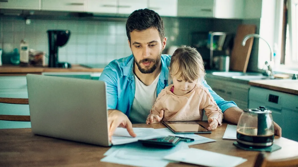 Man working on a laptop with a young child on his lap at a kitchen table.