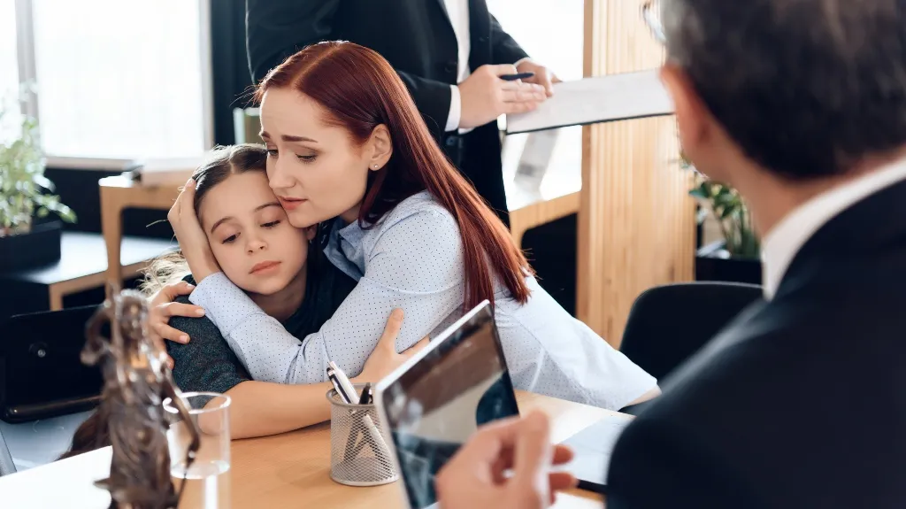 Woman in light blue shirt comforting young girl in grey sweater in office setting.