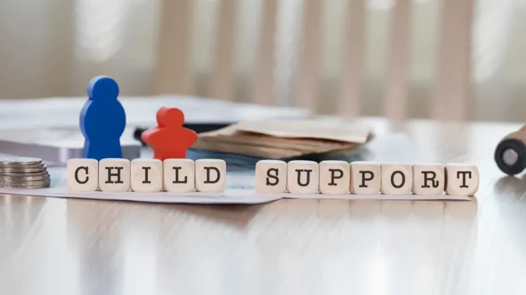 Wooden letter blocks spelling "CHILD SUPPORT" on a table with blue and red figurines, coins, and documents.