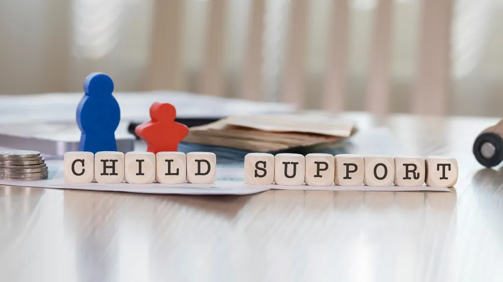 Wooden letter blocks spelling "CHILD SUPPORT" on a table with blue and red figurines, coins, and documents.