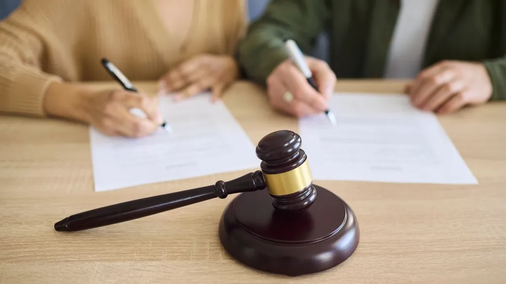 Wooden judge's gavel in foreground with two people signing documents in background.