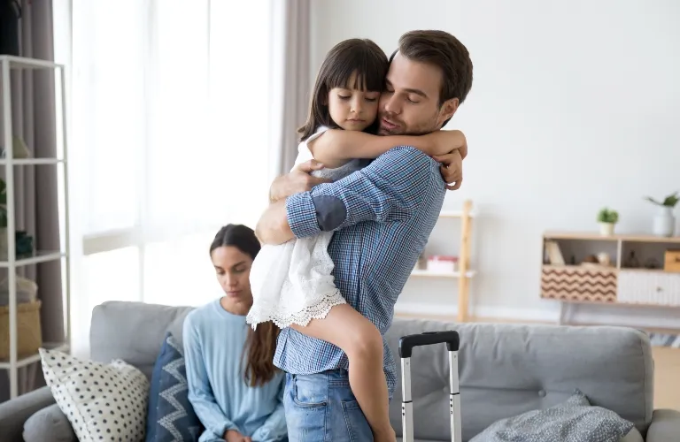 Man in blue checkered shirt holding young girl in white dress, woman seated on couch in background.