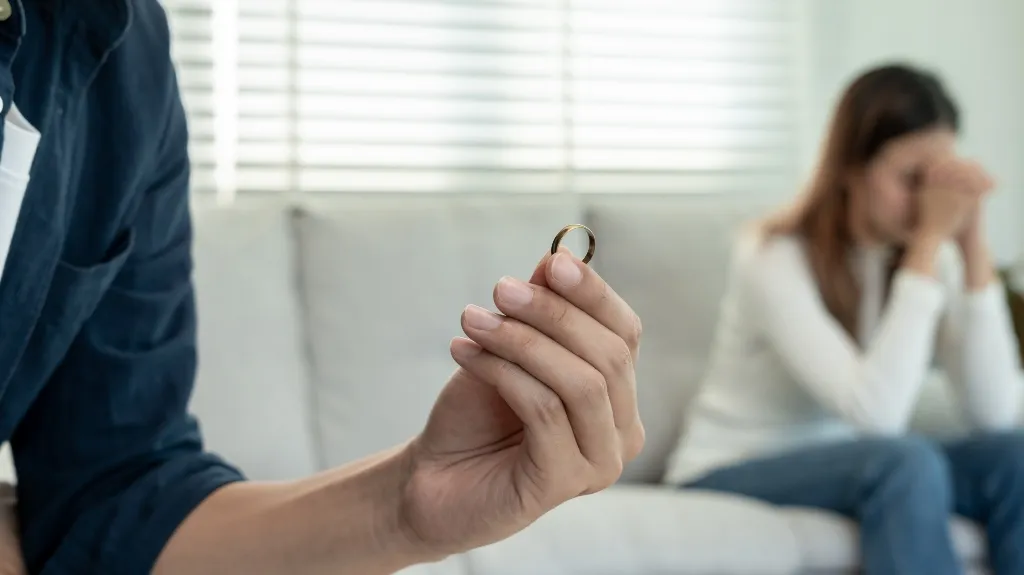 Close-up of a hand holding a wedding ring with a distressed woman sitting on a couch in the background.