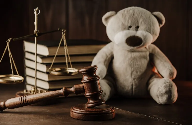 Wooden judge's gavel and scales of justice beside stacked books with a teddy bear in the background.