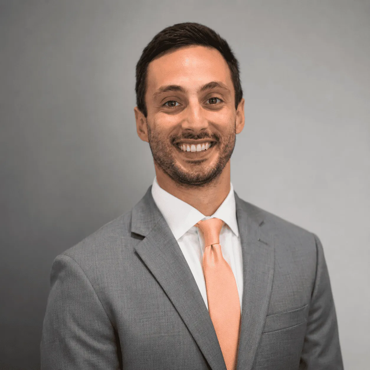 Professional headshot of a smiling man wearing a gray suit and peach tie against a neutral gray background.